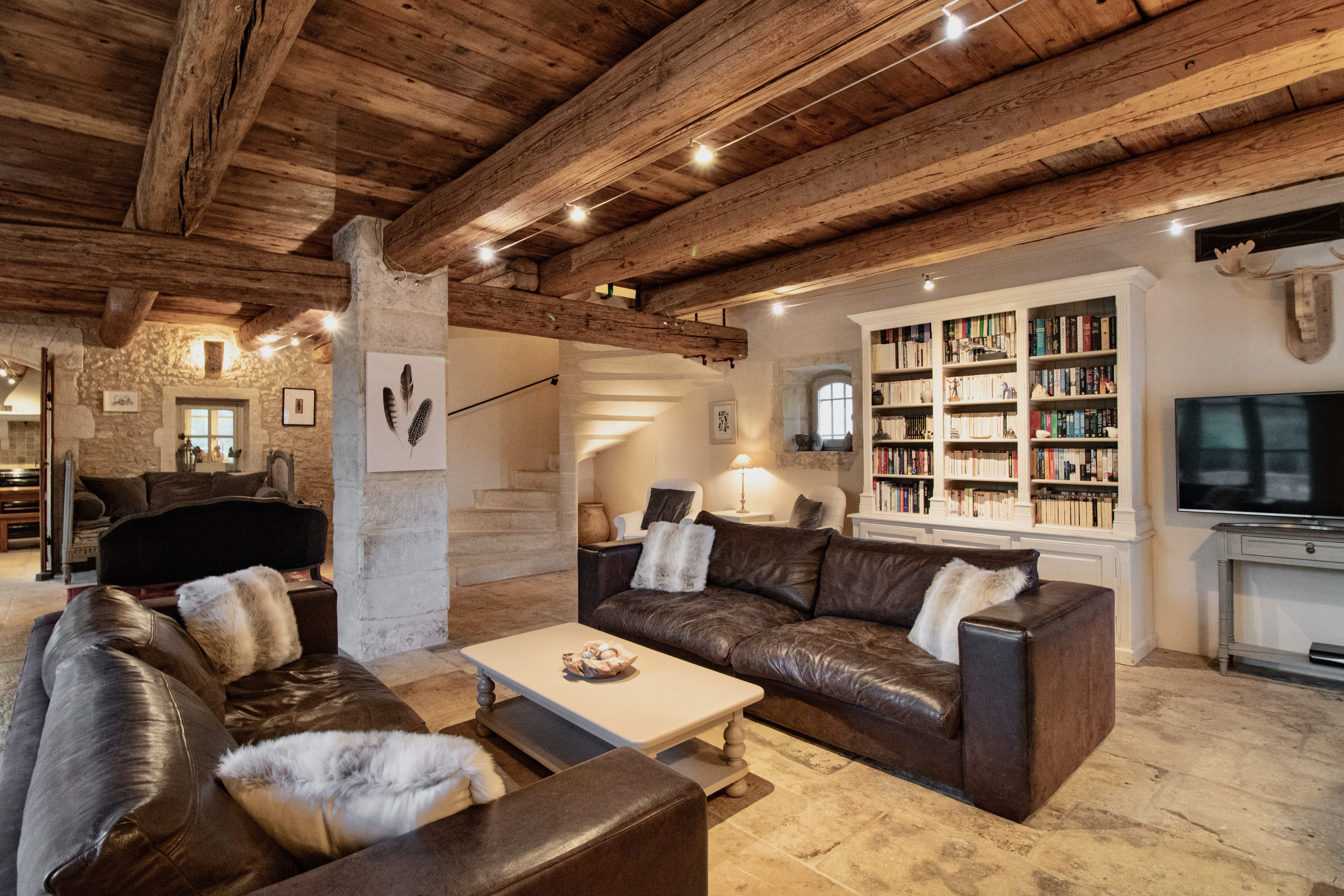 Cozy living room in a rustic home with exposed wooden beams, a stone pillar, a large leather sofa, fluffy chairs, a well-stocked bookshelf, and a modern tv.